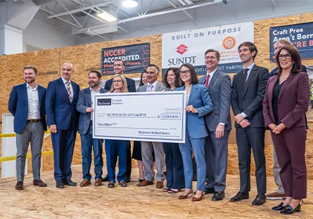 A group of professionals in business attire stand together in a construction training facility, smiling and holding an oversized check for $3 million from the Blackstone Charitable Foundation. Signs in the background display “NCCER Accredited” and “Sundt Construction.”