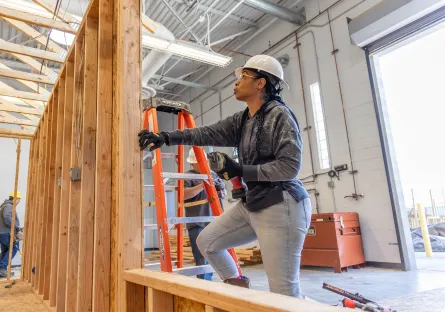 A woman wearing a white hard hat and safety gloves works on a wooden wall frame inside a construction training facility. She is using tools while standing on a platform, with ladders and other workers visible in the background.