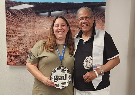 Rio Salado College graduate Kim Smock stands beside Lauren, a staff member, smiling for a photo indoors. Kim wears a white “Veteran” stole and black shirt, while Lauren holds a round “Congrats Grad” plate. A scenic desert landscape photo hangs on the wall behind them, celebrating Kim’s academic achievement.