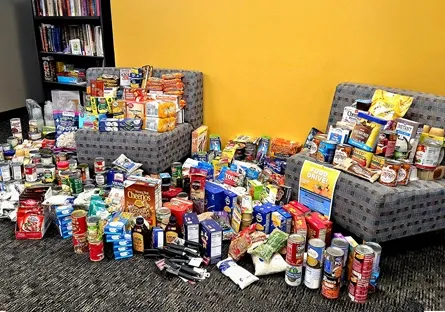 Large assortment of donated food items, including canned goods, boxed meals, snacks, and toiletries, displayed on the floor and chairs inside a building with a yellow wall.