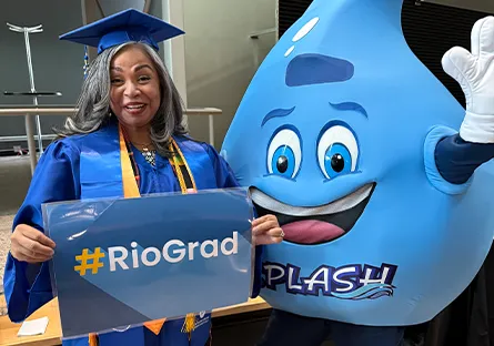 Magdalena, A Rio Salado College graduate in a blue cap and gown smiles while holding a '#RioGrad' sign. She stands next to Splash, the college’s blue water-drop mascot, who is waving and posing for the photo.