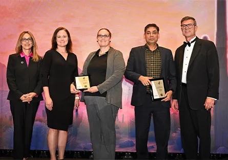 Five people stand in a row on a stage in front of a warm desert-themed backdrop. In the center, Amber Struthers holds an award plaque and smiles for the camera. Another award recipient stands to her right holding a similar plaque. Two presenters stand on the left, and one presenter in a black tuxedo stands on the far right.