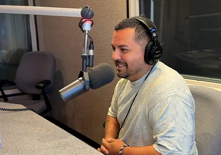 Frank Nino IV in the recording studio during his podcast interview. He is wearing a gray shirt and headphones, smiling.