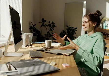 A young woman in a green hoodie sits at a desk in a sunlit home office, speaking and gesturing with her hands during a video call on a desktop computer. A notebook, keyboard, coffee mug, and tablet are arranged on the wooden desk, with houseplants and a mirror in the background.