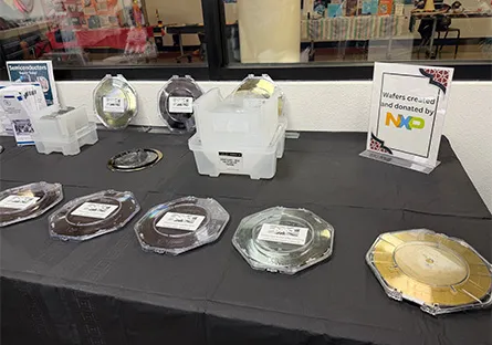 Display table showcasing semiconductor wafers in clear protective cases, with additional wafer containers in plastic bins behind them. A sign reads, ‘Wafers generously donated by NXP,’ and a classroom is visible through a window in the background.