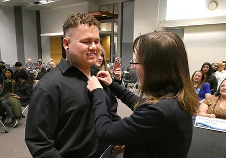 Yeison Ramirez Mendez smiles while a woman pins an item to his shirt during a ceremony as attendees watch from their seats.
