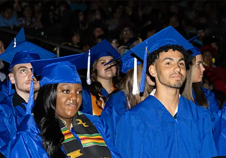 College graduates seated in an audience wearing blue caps and gowns, looking toward the stage during a ceremony.
