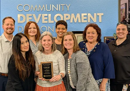 A group of professionals standing together indoors, one holding a plaque, in front of a wall with “Community Development” signage.