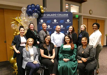 A group of staff members posing indoors in front of a Rio Salado College backdrop, with blue, gold, and white balloons arranged behind them.