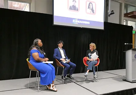 Three speakers seated on a stage during a panel discussion, holding microphones, with a presentation screen behind them.