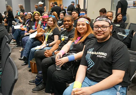 A row of participants wearing black “Riomagining” T-shirts, seated and smiling, some wearing colorful accessories and holding water bottles.