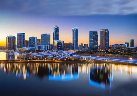 Downtown skyline at dusk with illuminated high-rise buildings and their reflections shimmering in the water along a riverfront.