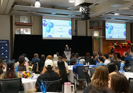 Audience seated at round tables watches a speaker at a podium during an ACE completion ceremony, with a large screen displaying “Congratulations ACE Completers” and balloons decorating the room.