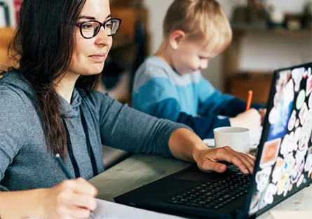 Adult learner working on a laptop at home while helping a child with schoolwork, illustrating flexible online learning in a real-life setting.