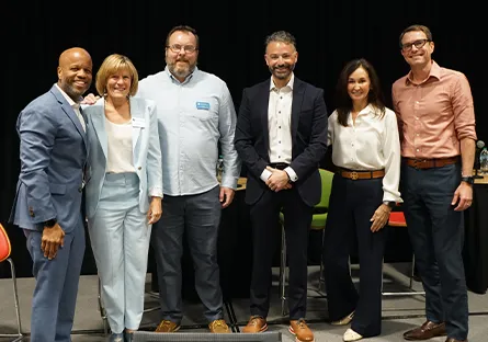 Six panelists stand together smiling in front of a stage during a professional event.