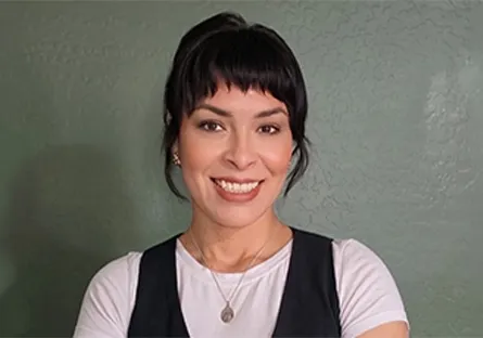 A woman with dark hair and bangs smiles at the camera while standing in front of a green wall, wearing a white shirt and black vest.