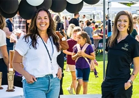 Two women smiling and standing under black and white balloons at an outdoor community or business event, with attendees gathered in the background.