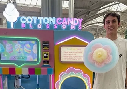 A young man stands beside a colorful “Cotton Candy Blossoms” vending machine while holding a large piece of pastel-colored cotton candy in a shopping mall.