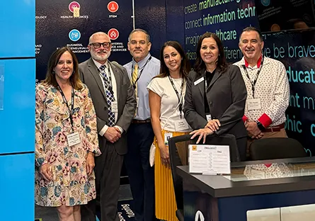 **Alt text:** Six professionals stand together smiling at a conference exhibit booth during an education and workforce event.