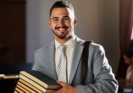 Smiling student in a light gray suit carrying a stack of books and a shoulder bag in an academic setting.