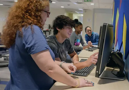 An instructor stands beside a student as they work together at a desktop computer in a classroom, while three other students use computers in the background, illustrating collaborative, technology-enabled learning.