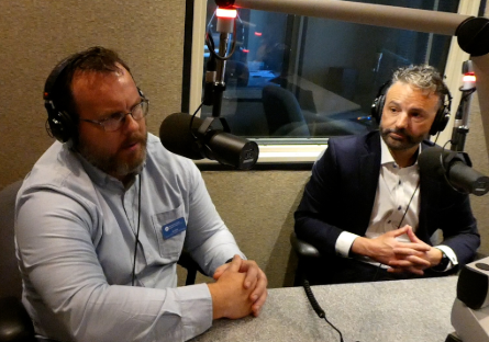 Ken Wilk and Zach Mannheimer wearing headphones speak into microphones during a podcast recording in a radio studio.