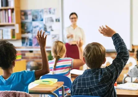 Students raise their hands to participate in a classroom discussion while a teacher stands at the front of the room.