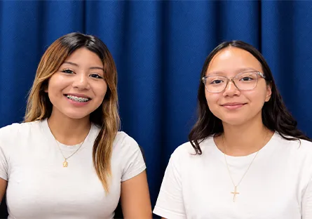 Allyson Hurtado Fernandez, left, and Silvana Castro Aguilar, right, smile at the camera while seated together in front of a blue curtain.