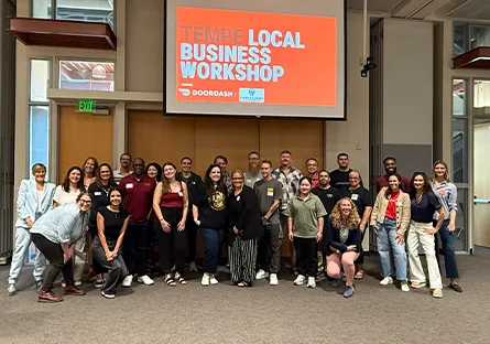 Attendees of the Tempe Local Business Workshop pose for a group photo beneath a presentation screen displaying the workshop name and DoorDash logo.