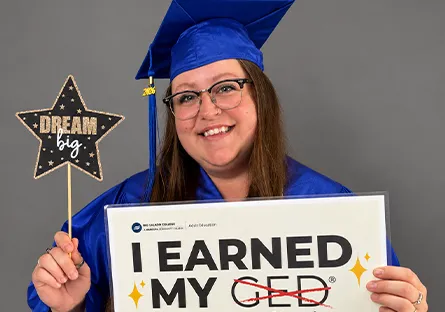 Brianna Reynolds smiles in a blue graduation cap and gown while holding a star-shaped “Dream Big” sign and a sign reading, “I earned my high school equivalency diploma.”