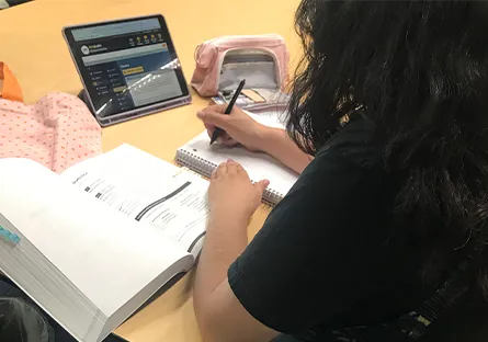 Rio Salado College student Dani Hernandez studies at a table, taking notes in a workbook while using a laptop.