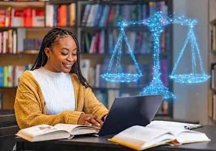 A smiling woman with long braids wearing a mustard-yellow cardigan works on a laptop at a library table with open textbooks. A glowing blue digital illustration of the scales of justice appears above the laptop, representing legal studies and online education.