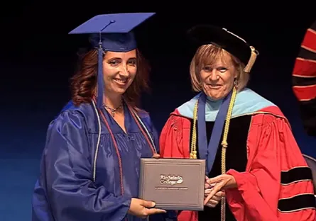 Diana Loya, wearing a blue graduation cap and gown, smiles while receiving her Rio Salado College diploma cover from President Kate Smith dressed in red academic regalia during the commencement ceremony.