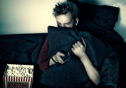 Scared teen hiding behind pillow sits on couch with bucket of popcorn, holding a remote
