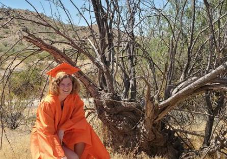 photo of Emma Harlow in a desert landscape kneeling under a dead tree in her graduation regalia.