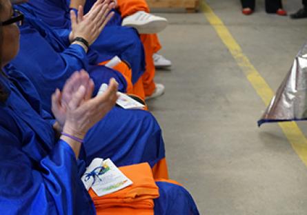 A group of individuals in blue graduation gowns and orange pants are seated in a row, clapping. The focus is on their hands and laps, with one person holding a colorful program booklet and folded orange fabric. The scene is part of a graduation ceremony taking place at Arizona State Prison Complex-Perryville