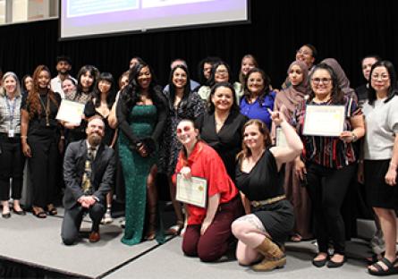 A large, diverse group of students and staff pose together on a stage at a Rio Salado College event, celebrating achievements. Several individuals proudly hold certificates, while others smile toward the camera. A woman in a sparkling green gown stands near the center. Behind the group, a projection screen displays a photo and testimonial from a student named Karen “Kiki” along with her written message.