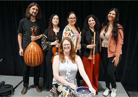 ASU’s Middle East Music Ensemble, Layali Al-Sham, pose together on a stage in front of a black curtain. Three women hold traditional Middle Eastern instruments, including a ney (flute) and a darbuka (goblet drum), while a man on the left holds an oud (lute). The seated woman in front smiles while resting her hands on the darbuka. 
