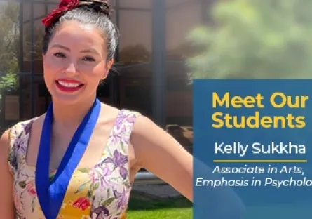Kelly Sukkha, a Rio Salado College student, smiles outside in the sunlight wearing a floral dress and a blue medallion. A graphic overlay reads: “Meet Our Students – Kelly Sukkha, Associate in Arts, Emphasis in Psychology.” The background shows a modern building with windows and greenery.