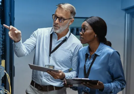 A man and a woman in professional attire stand in a server room, examining equipment. Both wear ID badges and hold tablets, collaborating while the man points at one of the servers. The environment is lit with a cool blue tone, emphasizing a high-tech workspace.