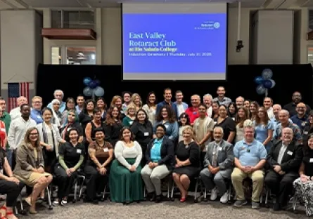 A large group of people pose together for a photo during the East Valley Rotaract Club charter celebration at Rio Salado College. The attendees, dressed in business and semi-formal attire, fill the stage area and front rows of the event space. Behind them, a projection screen displays the event title and date: “East Valley Rotaract Club at Rio Salado College, Charter Celebration, Thursday, July 25, 2025.” An American flag and balloon decorations are also visible in the background.