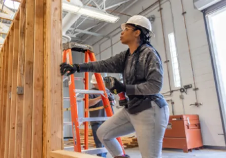 A woman wearing a white hard hat and safety gloves works on a wooden wall frame inside a construction training facility. She is using tools while standing on a platform, with ladders and other workers visible in the background.