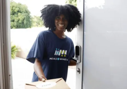 Smiling Meals on Wheels volunteer wearing a navy T-shirt delivers a boxed meal while standing at an open front door.