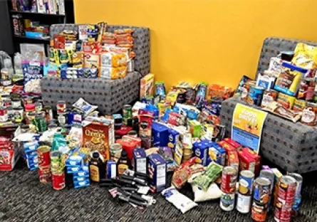 Large assortment of donated food items, including canned goods, boxed meals, snacks, and toiletries, displayed on the floor and chairs inside a building with a yellow wall.