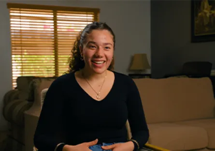 Alexandria Woodruff sits smiling on a couch in a living room, wearing a black long-sleeve top, speaking confidently during an interview for Rio Salado College’s spring campaign.