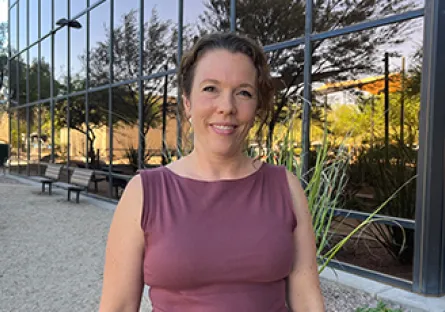 Alysa Call, with light brown hair pulled back stands outdoors in front of a reflective glass building. She is wearing a sleeveless mauve top and smiling at the camera, with desert landscaping and trees in the background.