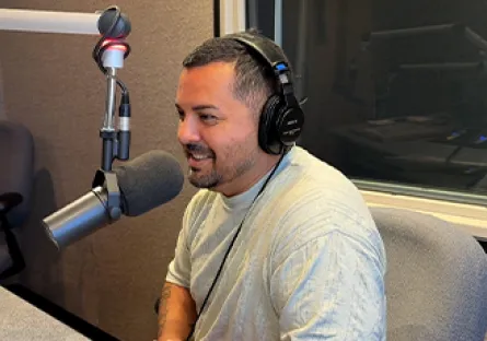 Frank Nino IV in the recording studio during his podcast interview. He is wearing a gray shirt and headphones, smiling.