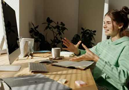 A young woman in a green hoodie sits at a desk in a sunlit home office, speaking and gesturing with her hands during a video call on a desktop computer. A notebook, keyboard, coffee mug, and tablet are arranged on the wooden desk, with houseplants and a mirror in the background.
