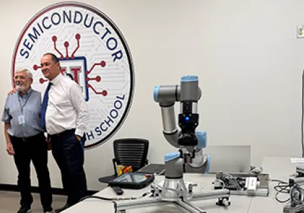 Two men stand together smiling in a classroom lab with a large ‘Semiconductor High School’ logo on the wall behind them. In the foreground, a robotic arm and electronic equipment sit on a worktable, highlighting the technology-focused environment.