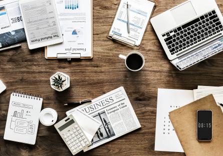 A cluttered desk with coffee cup, laptop newspaper, memos, and notebooks
